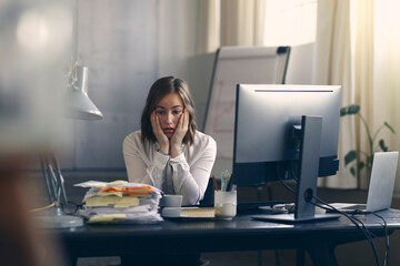 Very stressed business woman sitting in front of her computer looking at a large pile of paperwork - 354414451
