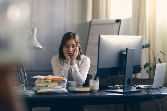 Very stressed business woman sitting in front of her computer looking at a large pile of paperwork