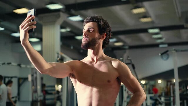 Handsome bearded young man with muscular wiry naked torso talking on video chat in modern gym, blogger writes content on mobile phone. Concept of healthy lifestyle. Shooting in slow motion.