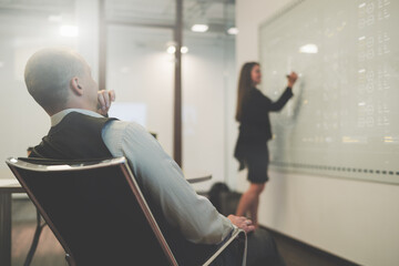 View from behind of a boardroom with a businessman in an office armchair closely observing how his female colleague in a defocused background makes a business presentation using electronic whiteboard