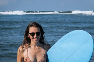 Happy smile young brunette woman hold blue surfboard going out of ocean after morning surfing. Portrait of beautiful girl spending summer holidays on the beach, enjoying sport, Bali, Indonesia, Asia