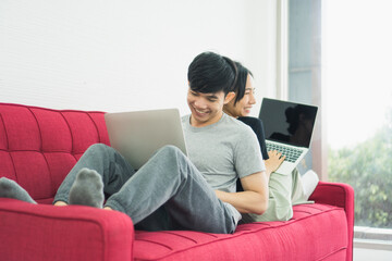 Young Asian couple love man and woman working at home by use computer laptop notebook sitting on red sofa in living room smiling and felling happy. corona virus, COVID-19 Pandemic new normal Concept