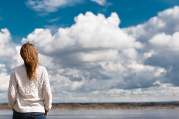 woman with red hair, dressed in a white sweater, stands with her back against the background of the river