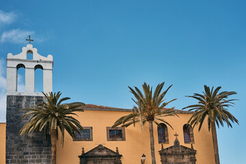 Fototapeta na wymiar Side view of the steeple and the yellow house wall of a Spanish church (Iglesia de Santa Ana) on Tenerife. In front of the church are three palm trees. The sky is blue and offers copy space.