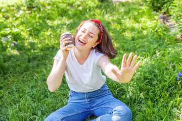 Fototapeta premium Happy girl smiling outdoor having lunch break. Beautiful young brunete woman with take away coffee cup resting on park or garden green grass background. Education freelancer resting leisure concept.