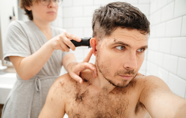 Fototapeta premium Portrait of a handsome man with a beard to whom. His wife cuts his head with a trimer in the bathroom. Haircut at home. Relationships in the family.