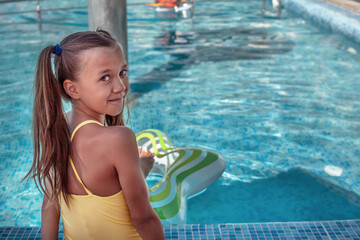 Back view of preteen girl dressed yellow swimsuit sitting in the pool and going to swim in the blue resort pool on the inflatanle ring. Summer vacation concept