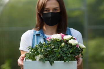Pretty woman in a flower greenhouse in a black protective mask holds a box with roses. We are safe. Coronavirus protection. Contactless flower delivery concept.