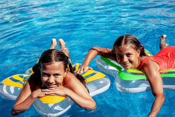 Two tween girls relaxing, laughing and have fun in the blue water of swimming pool with inflatanle...