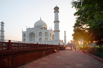 Fototapeta na wymiar A view of the Taj Mahal, Agra, at sunrise, with a path and trees. There is a wall and railing on the left and three visible minarets