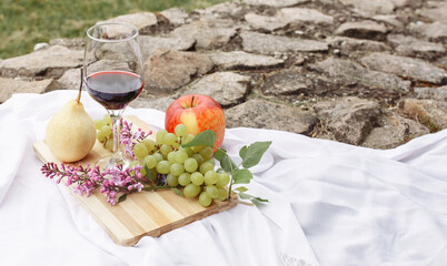 Picnic in the nature in the park. A glass of wine green grapes yellow pear red apple lilac on a wooden board on a white tablecloth in the grass in the garden on a stone path.
