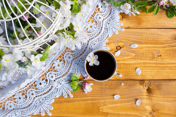 A cup of coffee with a fallen apple tree flower on a wooden table with a lace napkin. The metal cage is decorated with branches of a blossoming apple tree. Healthy breakfast concept. Interior concept.