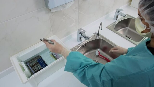 Wide shot of Doctor in white gloves wash dental medical instruments with a brush under a stream of running water from the tap. Before the disinfection procedure of orthodontic pliers.