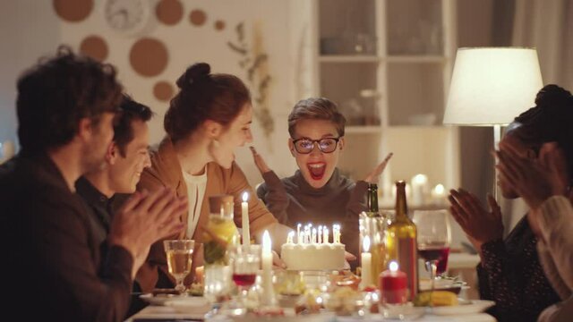 Young delightful Caucasian woman smiling and blowing candles on birthday cake while her cheerful multiethnic friends clapping hands at dinner party at home