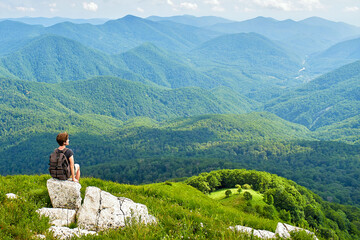 Woman traveler sits on top of a mountain with a beautiful view of the valley. A young female tourist with a backpack on top of a mountain.