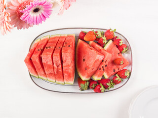 Watermelon slices with strawberries on ceramic tray on white wooden table with pink flowers. Fresh summer snack