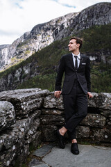 The groom in a black suit, standing at the edge of a stone wall against the background of mountains