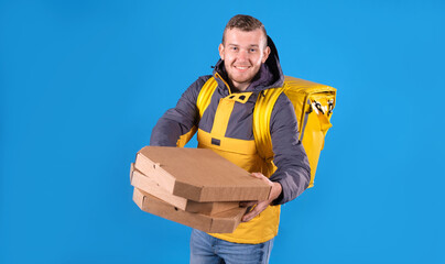 Happy food delivery man holds pizzas in cardboard boxes and holds them out for customer, dressed in yellow uniform and refrigerator bag on his back on blue background. Courier food delivery to home