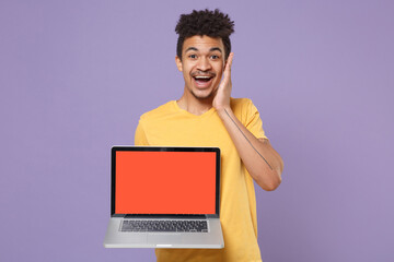 Excited young african american guy in yellow t-shirt isolated on violet background. People lifestyle concept. Mock up copy space. Hold laptop pc computer with blank empty screen, put hand on cheek.