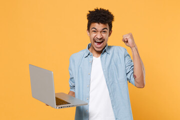 Screaming young african american guy in casual blue shirt posing isolated on yellow wall background studio. People lifestyle concept. Mock up copy space. Hold laptop pc computer, doing winner gesture.