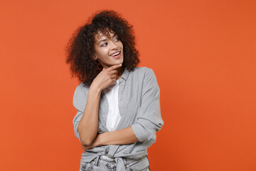 Cheerful young african american woman girl in gray casual clothes isolated on orange background studio portrait. People lifestyle concept. Mock up copy space. Put hand prop up on chin, looking aside.
