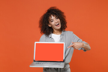 Naklejka premium Funny young african american woman in gray casual clothes isolated on orange background. People lifestyle concept. Mock up copy space. Point index finger on laptop pc computer with blank empty screen.