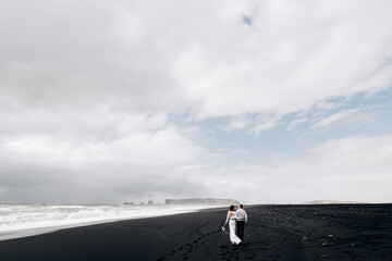 Destination Iceland wedding. A wedding couple is walking along the black beach of Vic. Sandy beach with black sand on the shores of the Atlantic Ocean. Huge foamy waves. The groom hugs the bride.