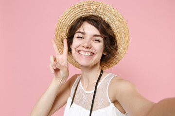 Close up of smiling tourist woman in dress hat isolated on pink background. Traveling to travel weekends getaway. Air flight journey concept. Doing selfie shot on mobile phone showing victory sign.