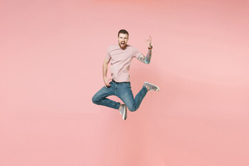 Fototapeta na wymiar Crazy young bearded tattooed man guy in pastel casual t-shirt posing isolated on pink wall background studio. People lifestyle concept. Mock up copy space. Jumping, holding hands like playing guitar.