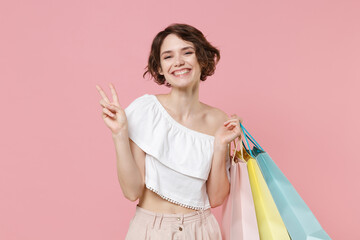 Smiling young woman girl in summer clothes hold package bag with purchases isolated on pastel pink background studio portrait. Shopping discount sale concept. Mock up copy space. Showing victory sign.
