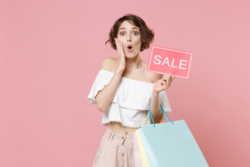 Shocked young woman girl in summer clothes isolated on pink background. Shopping discount sale concept. Mock up copy space. Hold package bag with purchases, sign with SALE title, put hand on cheek.