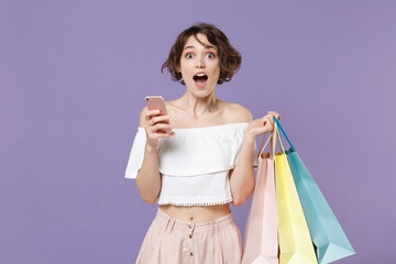 Shocked young woman girl in summer clothes hold package bag with purchases isolated on violet background. Shopping discount sale concept. Mock up copy space. Using mobile phone, typing sms message.