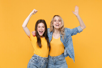 Two happy young women girls friends in casual t-shirts denim clothes posing isolated on yellow background studio portrait. People lifestyle concept. Mock up copy space. Clenching fists like winner.