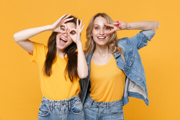 Two funny young women girls friends in casual t-shirts denim clothes isolated on yellow background. People lifestyle concept. Showing victory sign hold hands near eyes imitating glasses or binoculars.