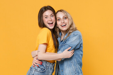 Two surprised young women girls friends in casual t-shirts denim clothes posing isolated on yellow...