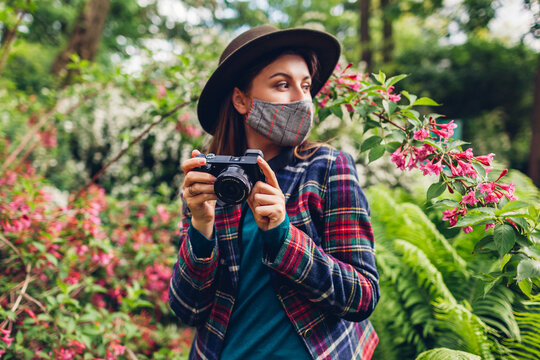 Woman photographer taking photos on digital camera in summer garden wearing coronavirus mask. Freelancer enjoys job