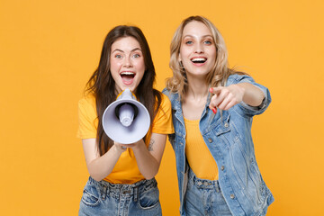 Two excited young women girls friends in casual t-shirts denim clothes isolated on yellow...