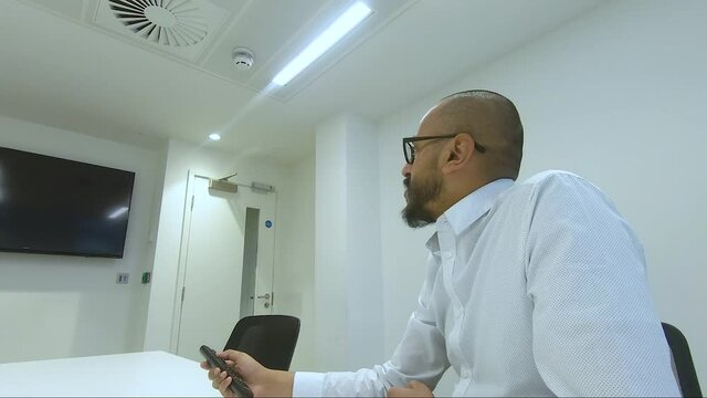 Adult UK Asian Male Sitting In Meeting Room Using Remote Control To Put TV On. Locked Off