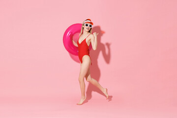 Beautiful young woman in red one-piece swimsuit cap glasses isolated on pink background. People summer vacation rest lifestyle concept. Mock up copy space. Hold swim inflatable ring point thumb aside.