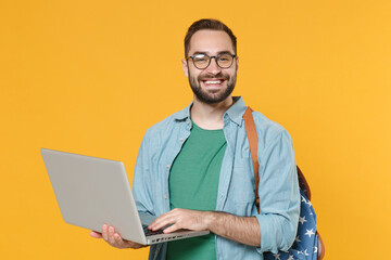 Smiling young man student in casual clothes glasses backpack isolated on yellow wall background. Education in high school university college concept. Mock up copy space. Working on laptop pc computer.