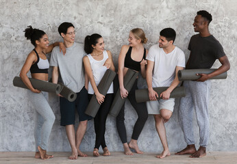 Fototapeta na wymiar Happy interracial friends in sportswear standing near wall with yoga mats