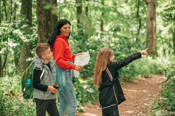 Beautiful mother with two kids hiking in a forest, looking at a map and pointing with hand.