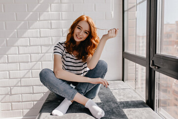 Elegant girl in jeans sitting on window sill and laughing. Emotional red-haired woman expressing happiness.