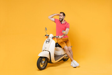 Fototapeta na wymiar Excited young bearded man guy in casual summer clothes standing near moped isolated on yellow background. Driving motorbike transportation concept. Holding hand at forehead looking far away distance.