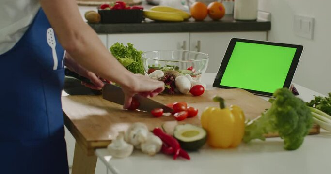The woman is slicing cherry tomatoes and looking at the tablet. Tablet with green screen. Salad preparation. There are lots of vegetables around. 4K