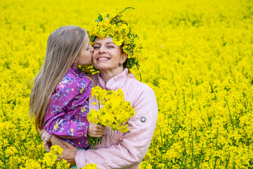 Cute girl with long light hair hug and kiss her mother in the field of yellow flowers. Rural landscape of blooming rapeseed. Mother's day concept. 