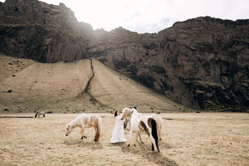 A bride stroking horses in a field against a rocky mountain. Destination Iceland wedding photo...