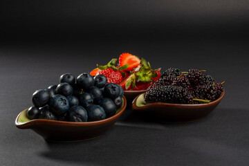 Close up on natural and organic plenty blueberries in front. Black mulberries in the middle and blurry red strawberries. Fresh fruits on dark background with shallow depth of field