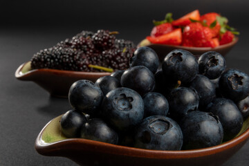 Very close up on natural and organic plenty blueberries in front. Black mulberries in the middle and blurry red strawberries. Fresh fruits on dark background with shallow depth of field