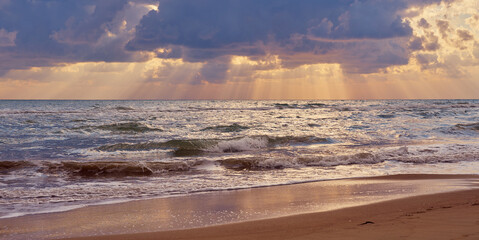 Obraz premium Colorful sunset at the tropical beach, sun behind clouds reflects on water and waves with foam hitting sand.